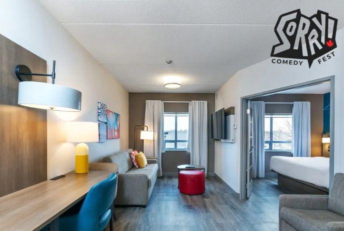 Modern suite with grey sofa red ottoman desk lamps and bed separated by open doorway with bright natural window light.