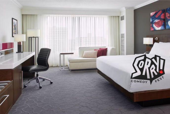 Modern hotel room with white bed, desk, sofa, lamps, and “SORRY! COMEDY FEST” logo.