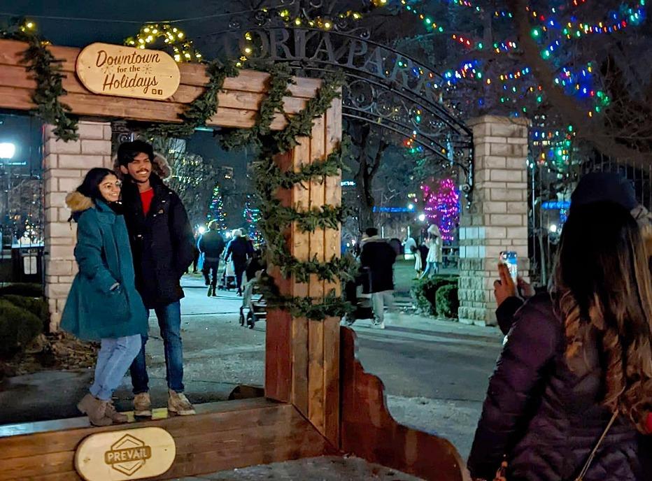 A couple posing for a photo under a sign that reads Downtown for the Holidays located in Victoria Park in London, Ontario