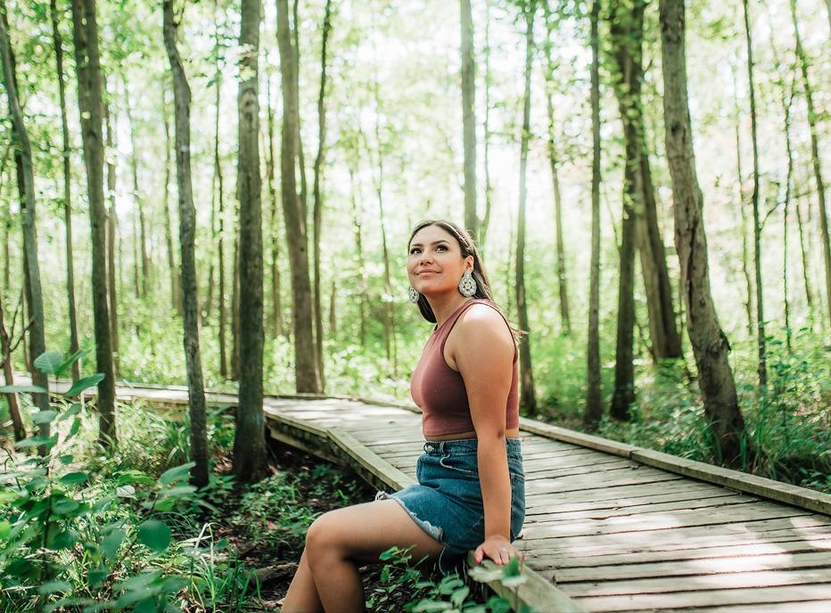 A female siting on the edge if a wooden path surrounded by tall trees in Sifton Bog, located in London, Ontario