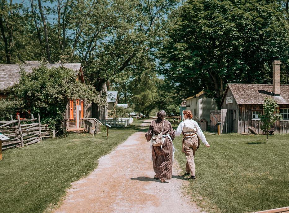 Two females walking down a dirt path by old wooden homes from the 1800s in Fanshawe Pioneer Village, located in London, ON
