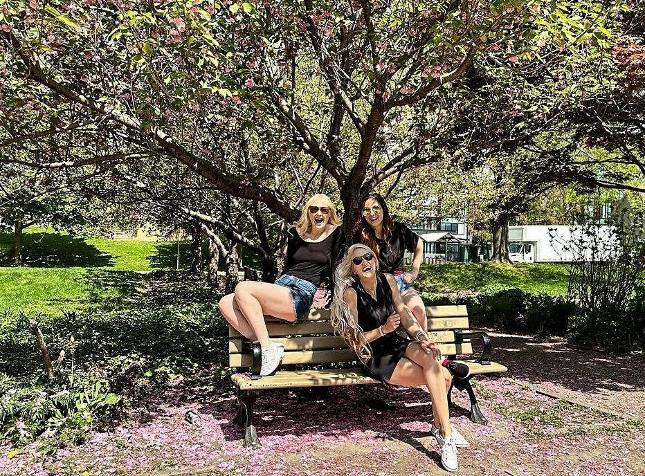 Three woman posing at a park bench outdoors with a large tree behind them and park, located in Wortley Village in London, ON