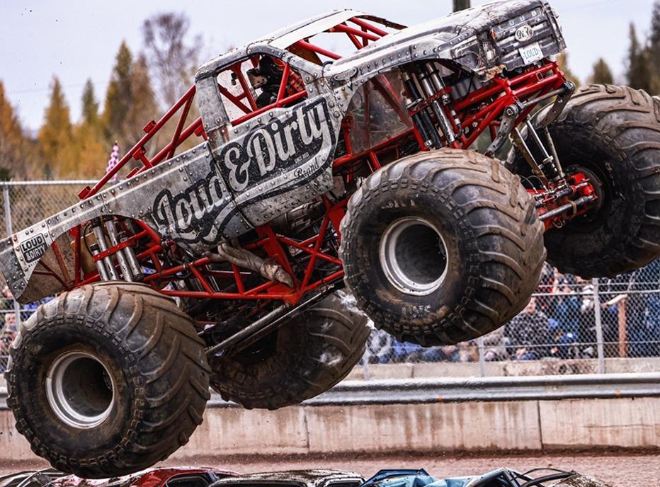 A monster truck in mid air, jumping over a group of crushed cars.