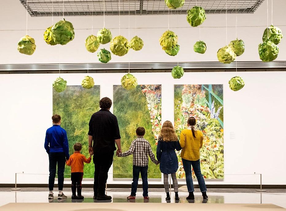 A family all holding hands, viewed from behind, looking at paintings on display at Museum London, located in London, Ontario