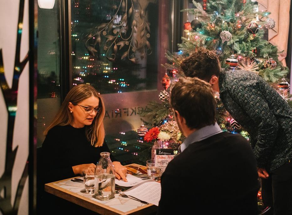 A couple dining at a restaurant with a server looking on as they order food with a Christmas tree behind them