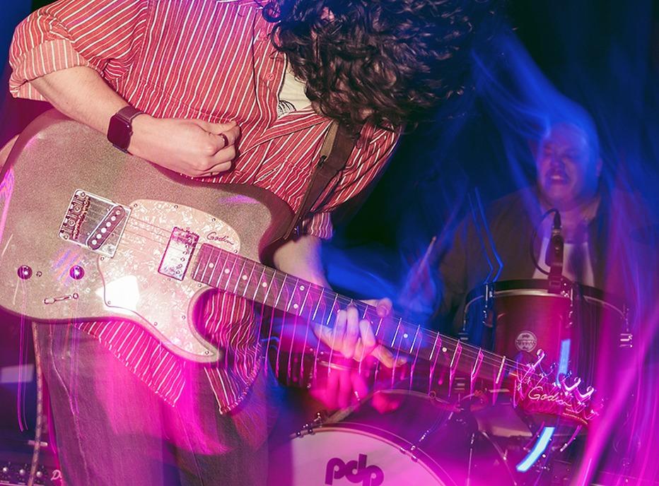 A band playing live on stage, the focus is on a musician playing the electric guitar and in the background is a person playing drums