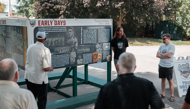 A group of people attending a tour of Labatt Park, The World's Oldest Operating Baseball Grounds and looking at a historic mural on display