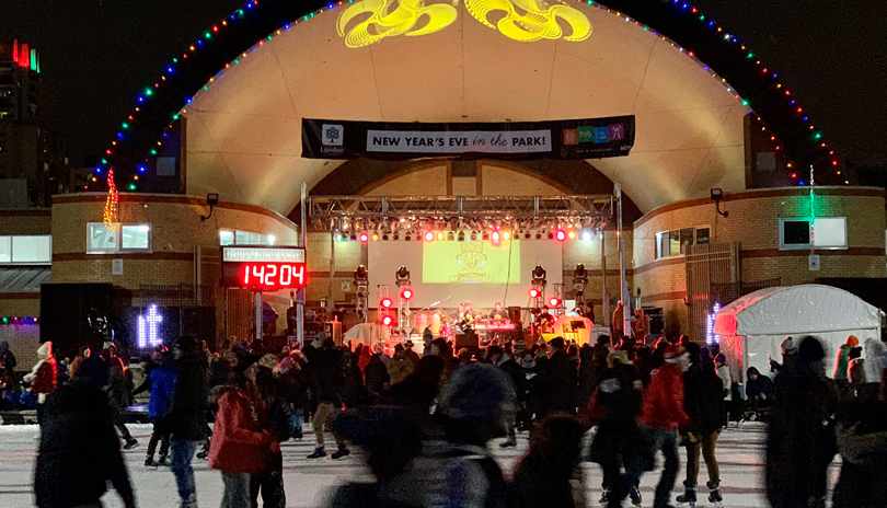 A live band playing in the bandshell outdoors in Victoria Park while people are skating on the outdoor ice rink, located in London, Ontario, Canada