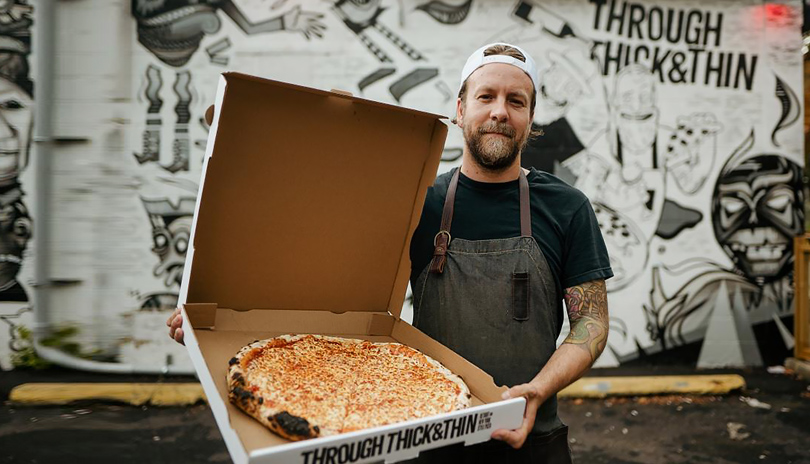 A chef holding an open box of pizza outside of a mural at Through Thick & Thin located in London, Ontario