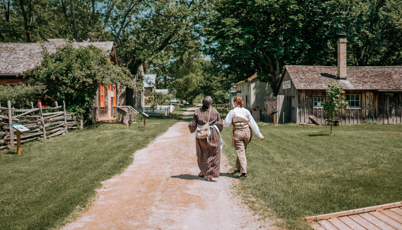 Two people walking down a path surrounded by historic buildings in Fanshawe Pioneer Village located in London, Ontario