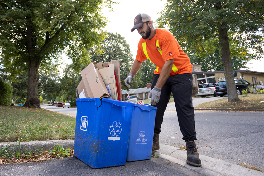 A city worker picking up a blue recycle box at the curb side of a house
