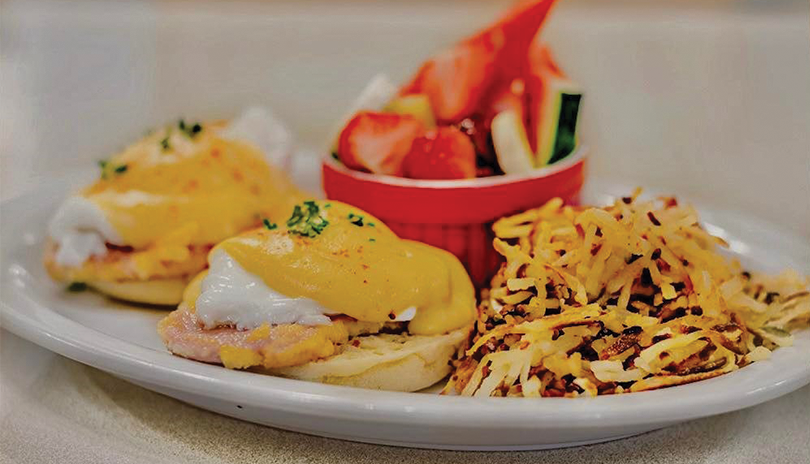 eggs benedict, shredded potato hashbrowns and fruit in a red dish displayed on a white plate