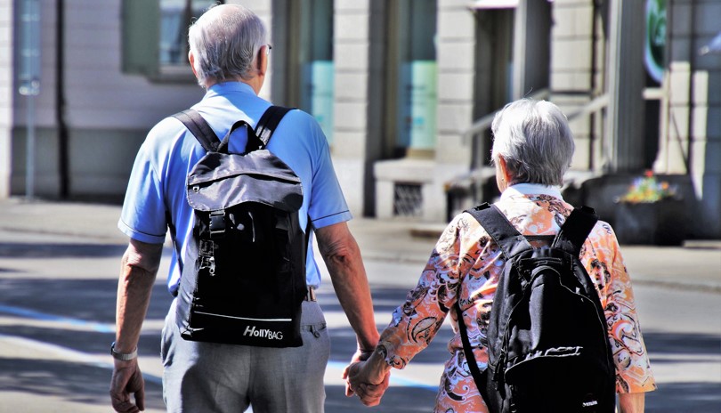 The backs of an elderly couple, a man and a woman, walking on a city sidewalk, holding hands and wearing backpacks