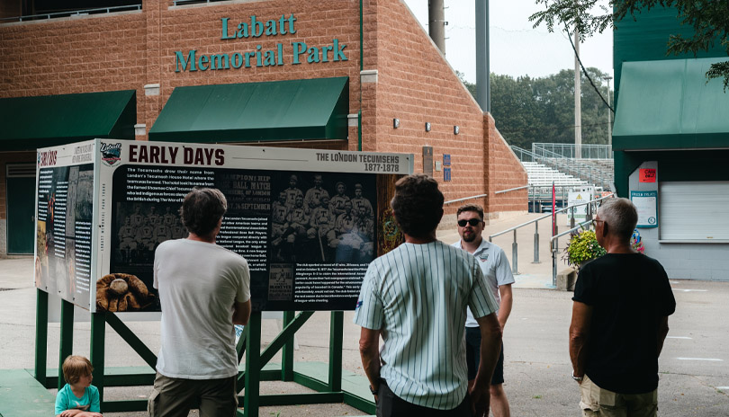 A group of people attending a tour of Labatt Park, The World's Oldest Operating Baseball Grounds and looking at a historic mural on display