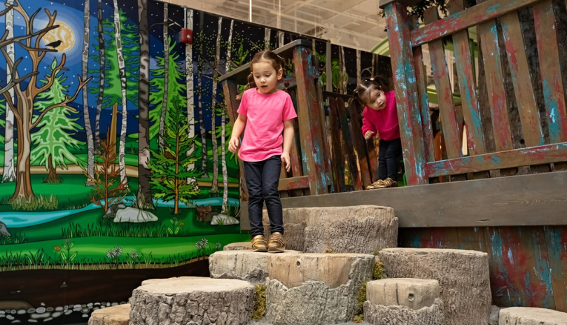 Two kids walk down stone steps in front of a mural at the London Children's Museum.