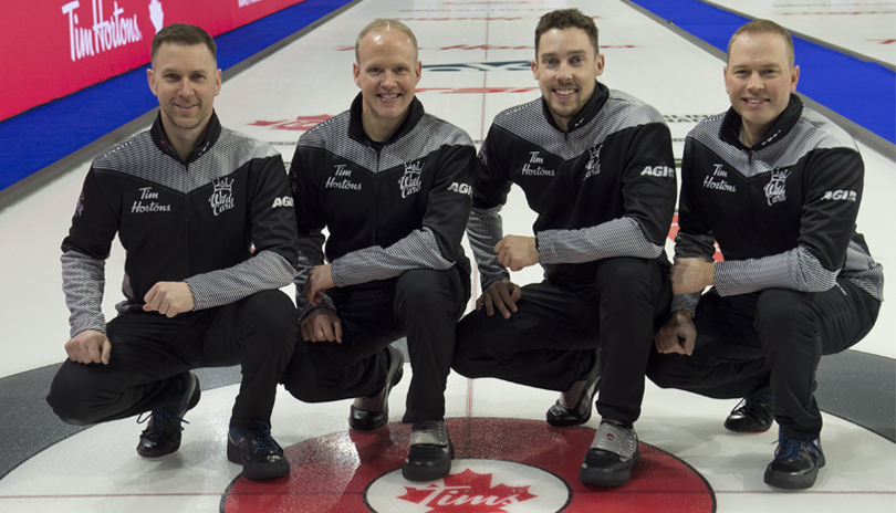 Four male curlers crouched and posing for a group shot on an ice rink.