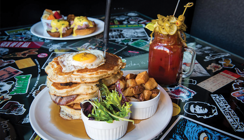 eggs benedict with potatoes and fruit on one plate, pancakes with a fried egg, salad and potatoes on a second plate with a caesar displayed on a table