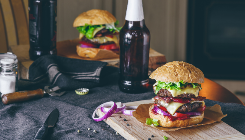 Two gourmet burgers resting on a wooden plate with two beers on the side 
