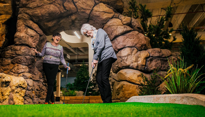 Two older women play mini putt on a Canadian themed course.