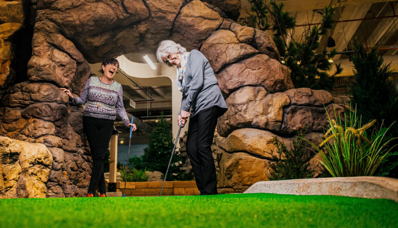 Two older women play mini golf under an archway of rocks at The Club House in London, Ontario.