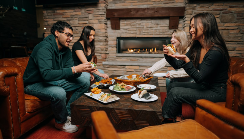 A group of friends eats from a full table of food by a fireplace.
