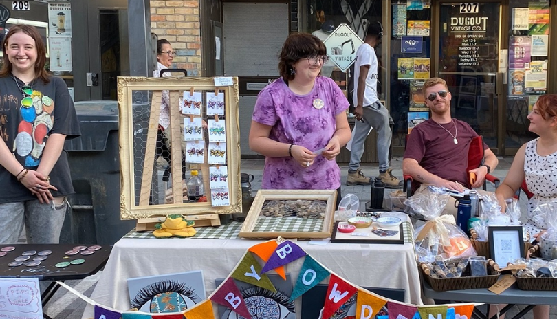 A group of artist vendors stand behind a colourful table on the street in London, Ontario.