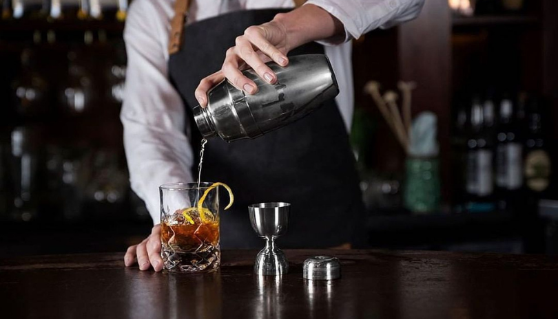 A bartender pouring a mixed drink into a glass with a lemon rind