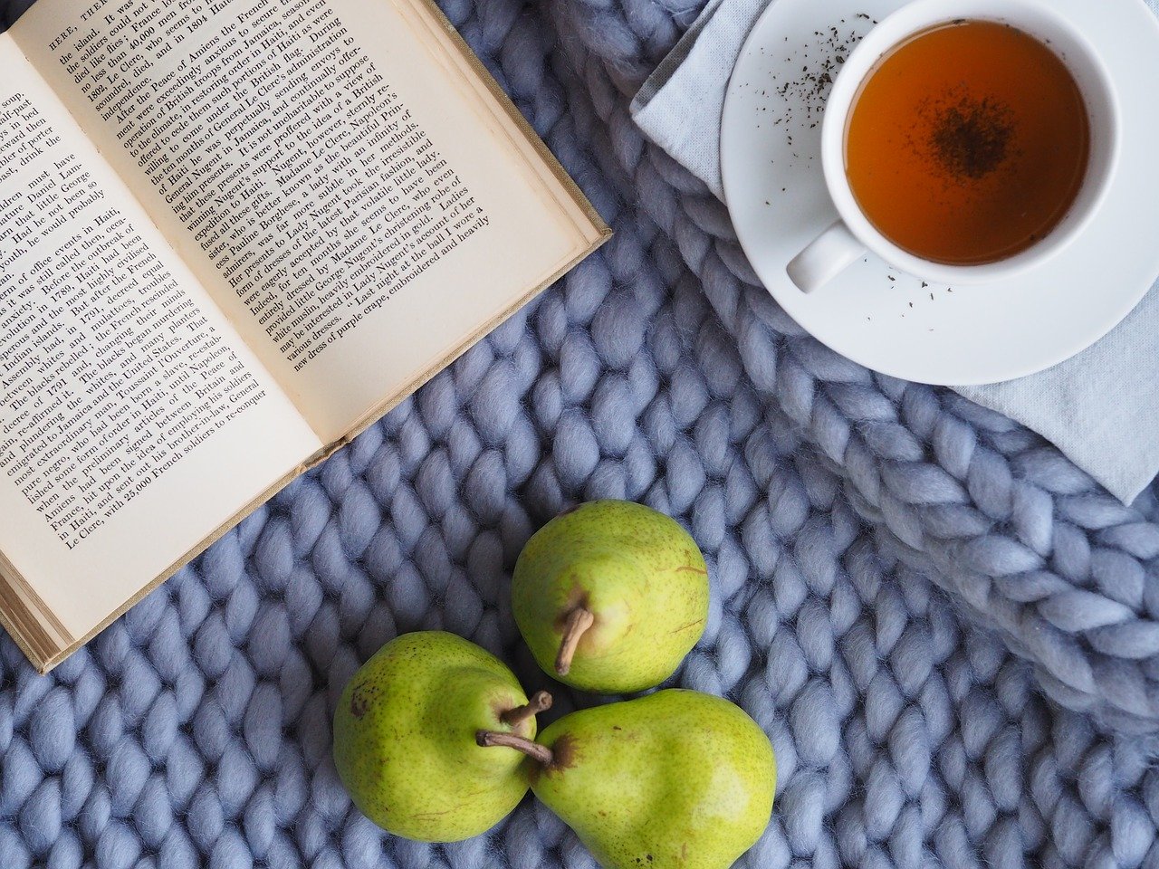 Table top view of an open book, a cup of tea and 3 pears, resting on a knitted blue blanket.