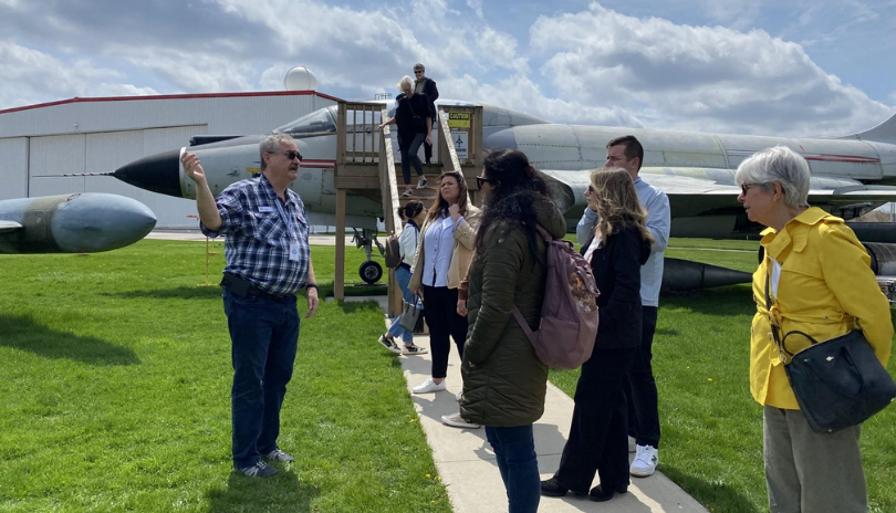 A tour group listens to a guide outdoors at the Jet Aircraft Museum with a plane in the background.