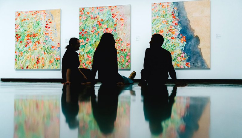 Three people sit on the floor of a gallery in front of a triptych of flower paintings.