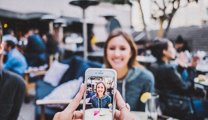 Person holding a cellphone in the foreground, taking a photo of a smiling woman seated at a table on a patio in the background