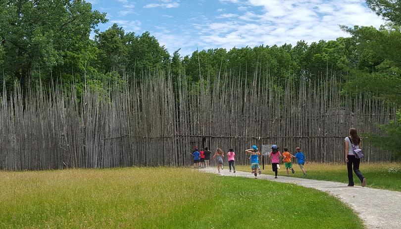 A small group of kids running into the entrance of wooden gates at Ska-Nah Doht Village's replica longhouse village