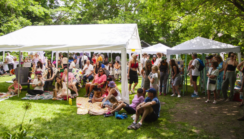 A crowd of people relaxed under white tents on a lawn surrounded by trees at Soft Fest in London, Ontario/