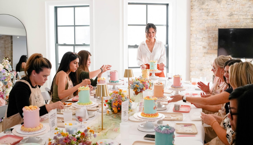 A group of women decorate cakes at a long table.