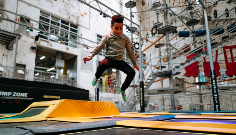 A kid jumps at an indoor trampoline park with a ropes course visible in the background.