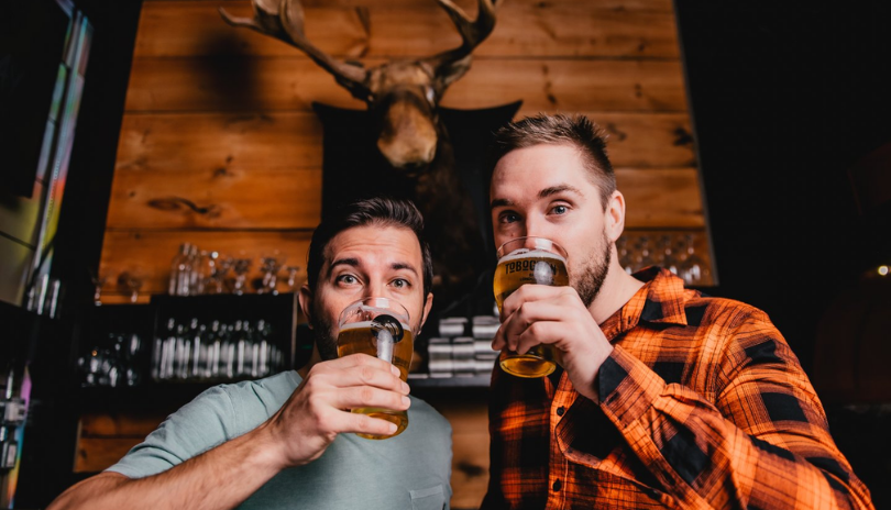 Two men sip beers in a rustic themed bar.