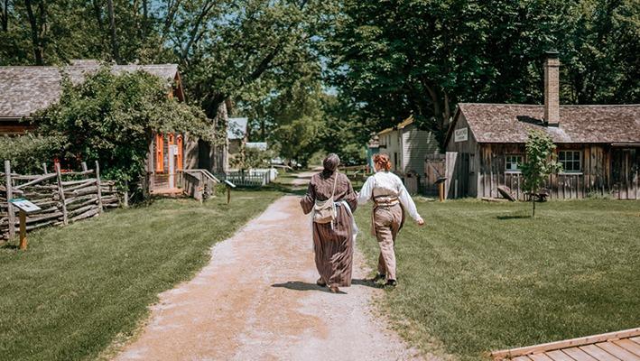 Two people walking down a dirt path with historic homes in the background at Fanshawe Pioneer Village located in London, Ontario