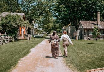 Two people walking down a dirt path with historic homes in the background at Fanshawe Pioneer Village located in London, Ontario