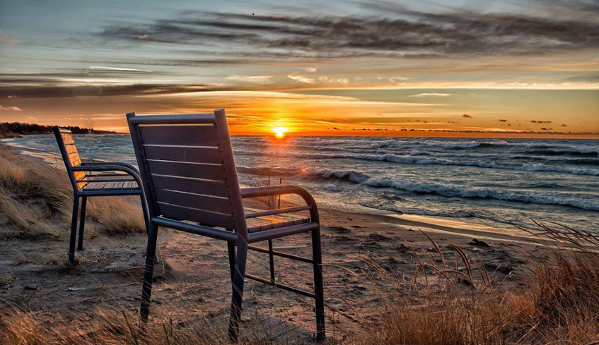 Two empty chairs near the shore of a body of water with the sun setting in the background and waves crashing in Kincardine, Ontario