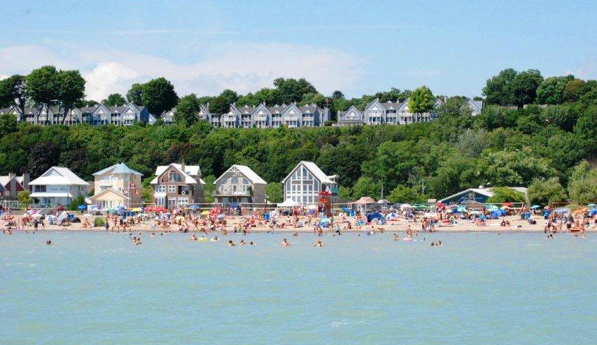 Large groups of people gathered at a beach in Port Stanley, Ontario with some in the water and some by the shore on a hot summer day.