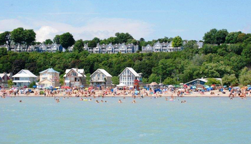 Large groups of people gathered at a beach in Port Stanley, Ontario with some in the water and some by the shore on a hot summer day.