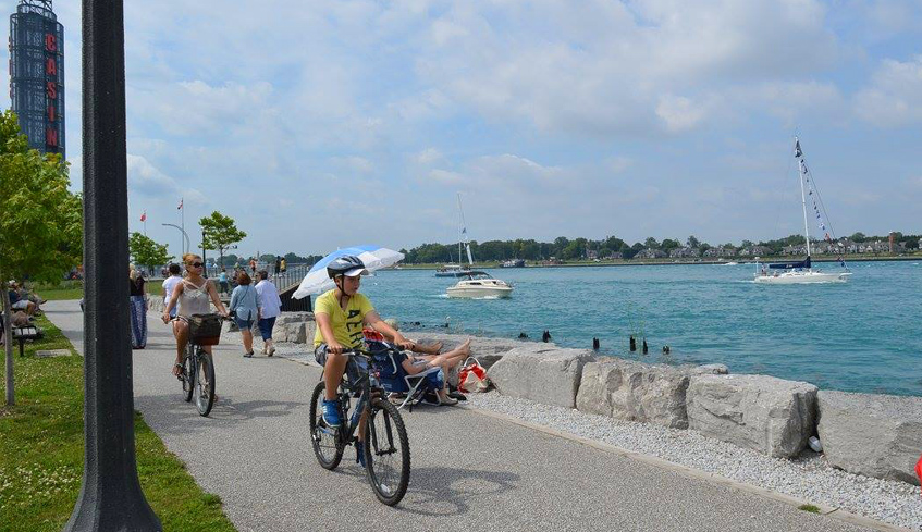 A boy and his mother on bikes riding on a paved path beside Lake Huron, with sailboats in the water in Sarnia-Lambton, Ontario
